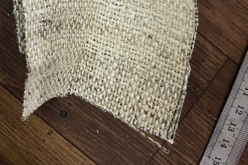 Close-up of a ruler-marked cut edge on handwoven hemp, a loose thread end trailing off one corner beside a steel ruler
