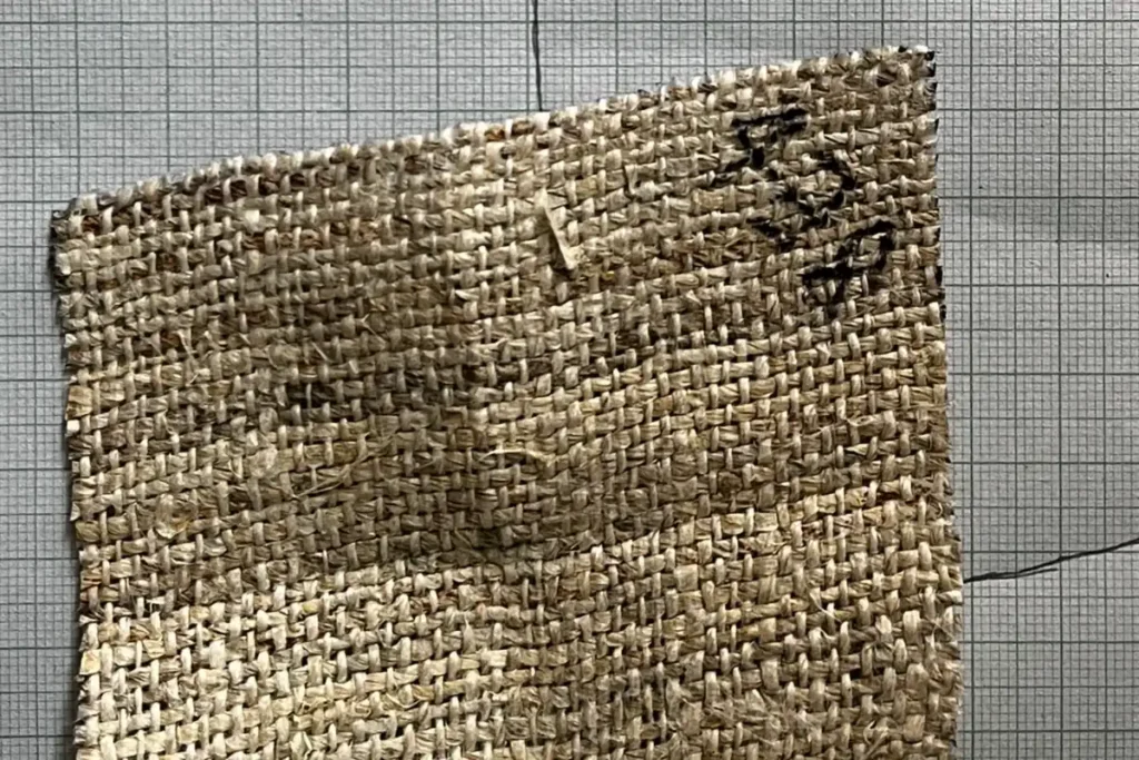 Close-up of a handwoven hemp swatch on graph paper, each edge trimmed along a single thread, the top edge slanting